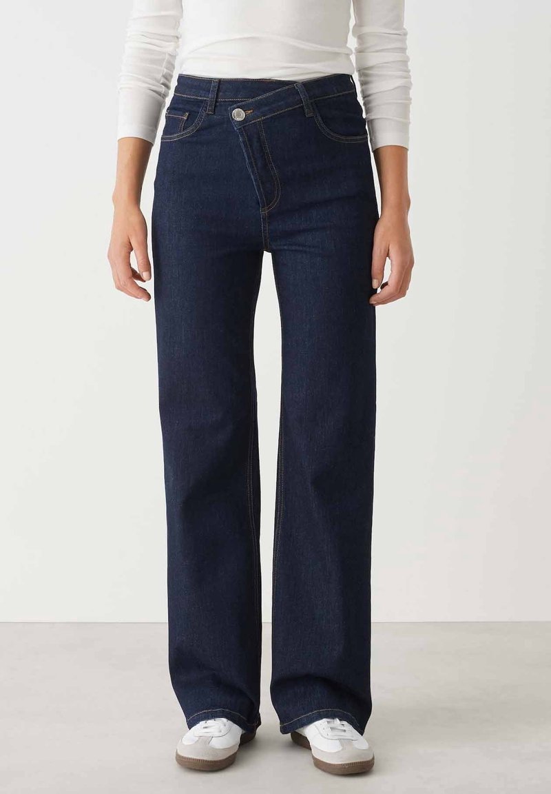 Woman wearing dark blue wide-leg jeans with an asymmetrical front button and white sneakers, standing against a plain background.