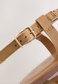 Tanfarvet lædersandal med en flettet rem og spænde i guld-tone. Glat sål med en let glans; justerbar pasform for komfort.