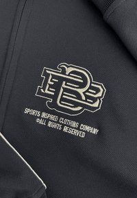 Zwarte stof met een geborduurd logo met gestileerde letters "PB." De tekst eronder luidt "SPORTGEÏNSPIREERD KLEDINGBEDRIJF © ALLE RECHTEN VOORBEHOUDEN."