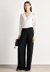 Γυναίκα που φοράει λευκή μπλούζα με V λαιμόκοψη και διαφανή μανίκια, μαύρο παντελόνι φαρδιάς γραμμής, μαύρα παπούτσια και κρατά μια μικρή μαύρη τσάντα.