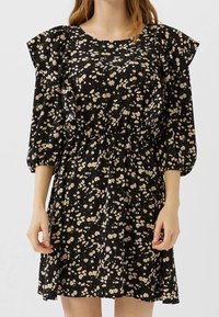 Robe fleurie noire en tissu léger, avec un col rond, des manches bouffantes, une taille élastique et de petits motifs floraux blancs et jaunes.