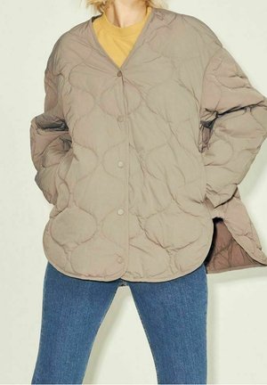 Persona con chaqueta beige acolchada abotonada sobre camisa amarilla y vaqueros azules, de pie con las manos en los bolsillos de la chaqueta.