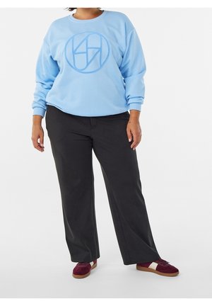 Persoon draagt een lichtblauwe trui met een ronde logo, zwarte broek met wijde pijpen, en bordeauxrode sneakers met witte strepen.