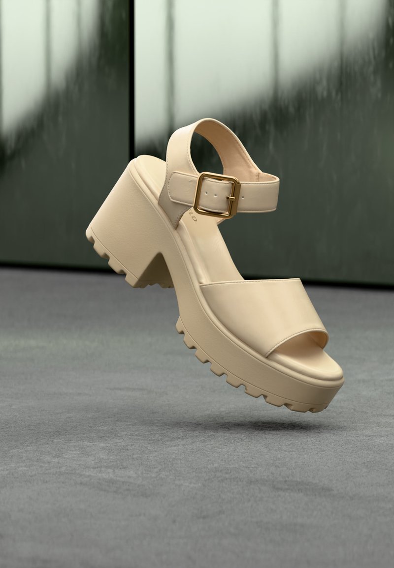 Μπεζ πλατφόρμα σανδάλι με χοντρή σόλα, ανοιχτό δάχτυλο, λουρί αστραγάλου και χρυσή αγκράφα, να αιωρείται πάνω από γκρι τσιμεντένιο πάτωμα.