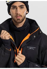 Giacca outdoor nera con accenti arancioni, chiusura zip frontale e logo SWEDEMOUNT. Il modello indossa una maglia base nera e un berretto coordinato.