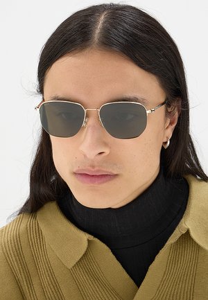 Persona con capelli lunghi e scuri che indossa occhiali da sole con montatura dorata, un dolcevita nero e un maglione a coste giallo senape con colletto.