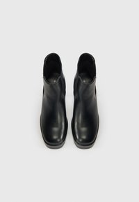 Botas de tobillo de cuero negro con un acabado suave, punta redondeada y laterales cortados. Tacón bajo de goma negra sólida. Diseño simplista.