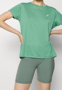 Camiseta deportiva verde claro con mangas cortas, cuello redondo y un pequeño logo. Combinada con pantalones cortos ajustados verdes a juego. Textura suave.