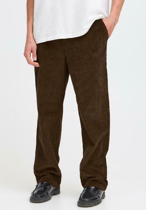 A person stands with one hand in their pocket, wearing a loose-fitting white shirt and brown corduroy pants, paired with black shoes.
