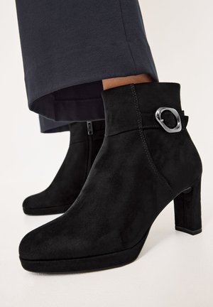 Schwarze Wildleder-Ankle Boots mit Blockabsatz, versehen mit einer silbernen Akzent-Schnalle und einer glatten Oberfläche, die ein elegantes Design präsentieren.