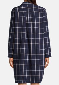 Una donna che indossa un abito lungo a maniche lunghe di colore blu navy con un motivo a quadri beige e azzurri, vista da dietro.