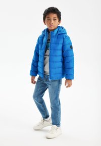 Blå pufferjacka med huva, med quiltad textur och elastiska manschetter. I kombination med ljusa jeanstyg och vita sneakers.