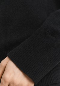 Hand in einem schwarzen, gerippten Strickpullover mit dezentem, gesticktem Logo nahe dem Handgelenk.