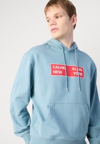 Mėlynas gobtuvėlis su priekiu kišene, kuriame yra raudoni tekstiniai langeliai su „CALVIN“ ir „KLEIN Y“ logotipais. Minkšta medvilnės mišinio medžiaga.