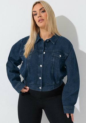 Lyshåret kvinde iført en mørkeblå denimjakke med sølvknapper og sorte bukser, stående mod en hvid baggrund.