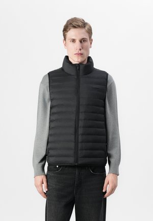 Zwarte gewatteerde gilet met hoge kraag, ritsluiting en horizontale gestikte ontwerpen, gelaagd over een grijze longsleeve shirt. Jeans maken de outfit compleet.