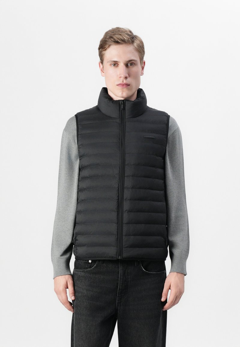 Zwarte gewatteerde gilet met hoge kraag, ritsluiting en horizontale gestikte ontwerpen, gelaagd over een grijze longsleeve shirt. Jeans maken de outfit compleet.