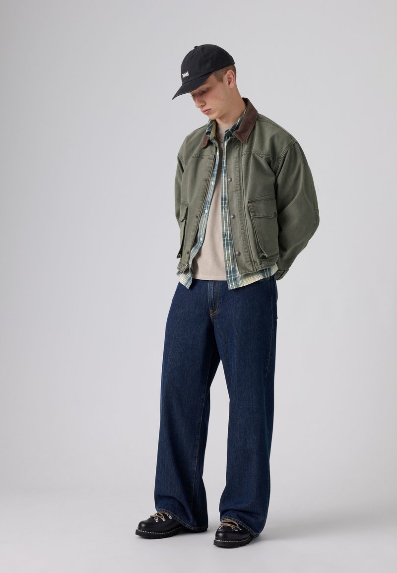 Jeune homme portant une veste verte, une chemise à carreaux, un t-shirt beige, un jean large bleu, une casquette noire et des chaussures noires, debout, tête baissée, les mains derrière le dos.