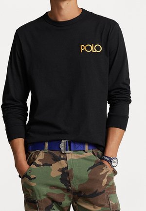 Person iført en sort langærmet skjorte med guld "POLO" logo, camouflagemønstrede cargobukser og et blåt bælte med spænde i sølv.