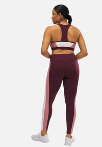 Sujetador deportivo burdeos y leggings con rayas laterales rosas y blancas. Los leggings cuentan con una cinturilla alta y una tela texturizada. El modelo lleva zapatillas blancas.