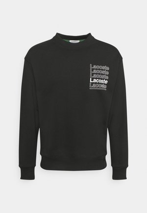 Zwarte lange mouwen sweater met ronde hals met herhaald witte "Lacoste"-tekst op de linkerborst, waarvan één vet en onderstreept.