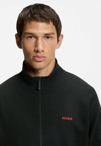 Giacca nera con zip in tessuto texture. Presenta un colletto alto e il logo rosso "HUGO" sul lato sinistro. Primo piano della parte superiore del corpo.