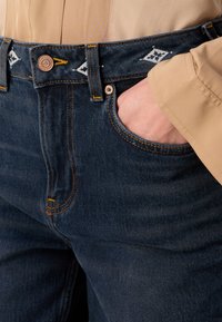 Mørk denimjeans med en strukturert passform, med en detaljert midjebånd med et logomønster, kobberfarge knapp og kontraststing.