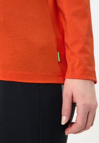 Langärmliges oranges Shirt aus Baumwolle mit einem seitlichen grünen Etikett, kombiniert mit schwarzen, eng anliegenden Hosen. Glatte Textur.