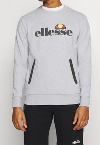 Hombre vistiendo un sudadera gris claro con el logo de "ellesse" y bolsillos con cremallera negros, combinado con pantalones cortos negros con un logo a juego.