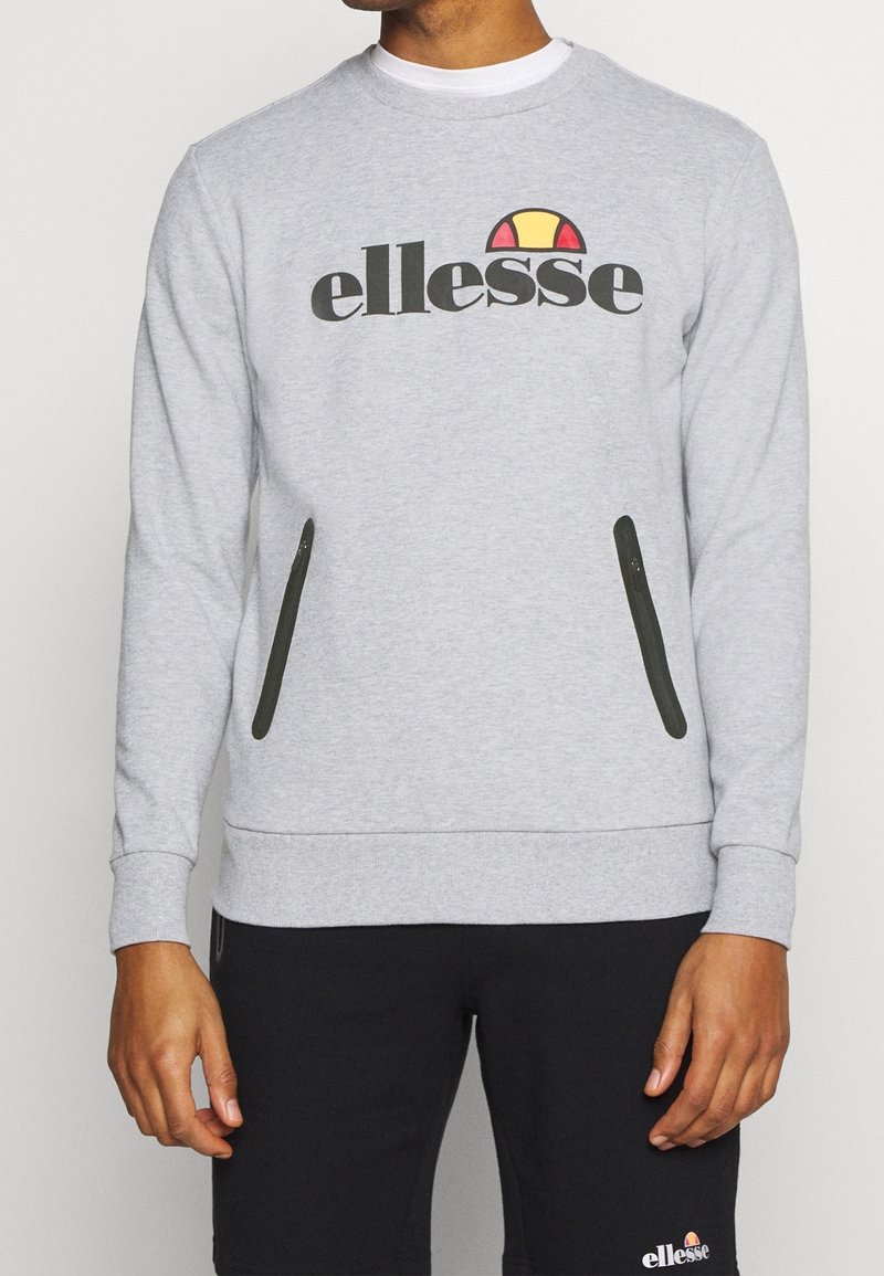 Hombre vistiendo un sudadera gris claro con el logo de "ellesse" y bolsillos con cremallera negros, combinado con pantalones cortos negros con un logo a juego.