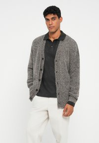 Grå strikket cardigan med en struktureret overflade, der har knapper foran og ribbede ærmekanter, båret over en sort polo og lyse bukser.