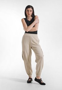 Beżowe spodnie cargo z elastyczną talią i mankietami, z bocznymi kieszeniami. Do tego czarna bezrękawnik i czarne wsuwane buty.