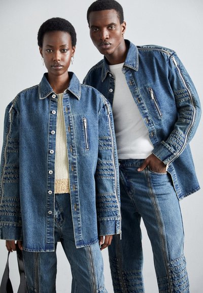 Jachete din denim și blugi de culoare albastru mediu, cu fermoare și accente din panouri texturate. Jachetele au buzunare mari; tricotajele sunt vizibile dedesubt.