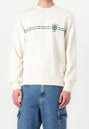 Homme portant un pull Lacoste blanc cassé à manches longues avec un logo de balle de tennis verte et des rayures sur la poitrine, associé à un jean cargo bleu.