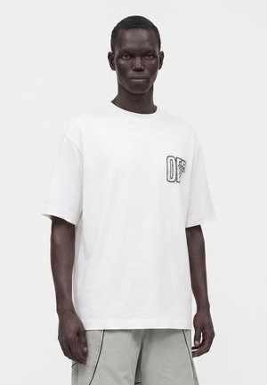 Modèle masculin à la peau foncée portant un t-shirt blanc ample à manches courtes avec un petit motif noir sur la poitrine et un pantalon gris clair.