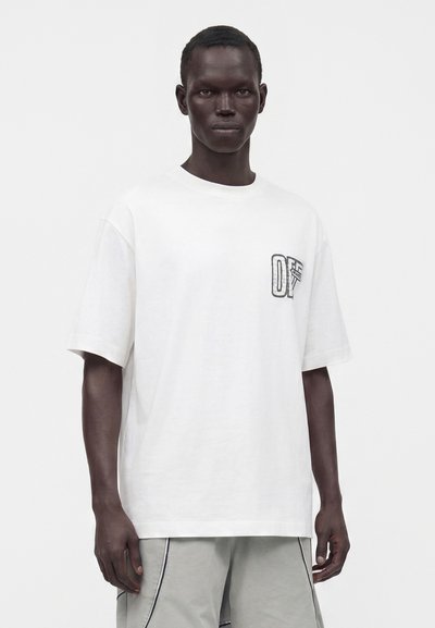 Modèle masculin à la peau foncée portant un t-shirt blanc ample à manches courtes avec un petit motif noir sur la poitrine et un pantalon gris clair.