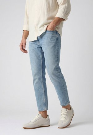 Mann trägt hellblaue Jeans, offwhite hochgekrempeltes Hemd und weiße Schnürsneaker, steht mit einer Hand in der Tasche.