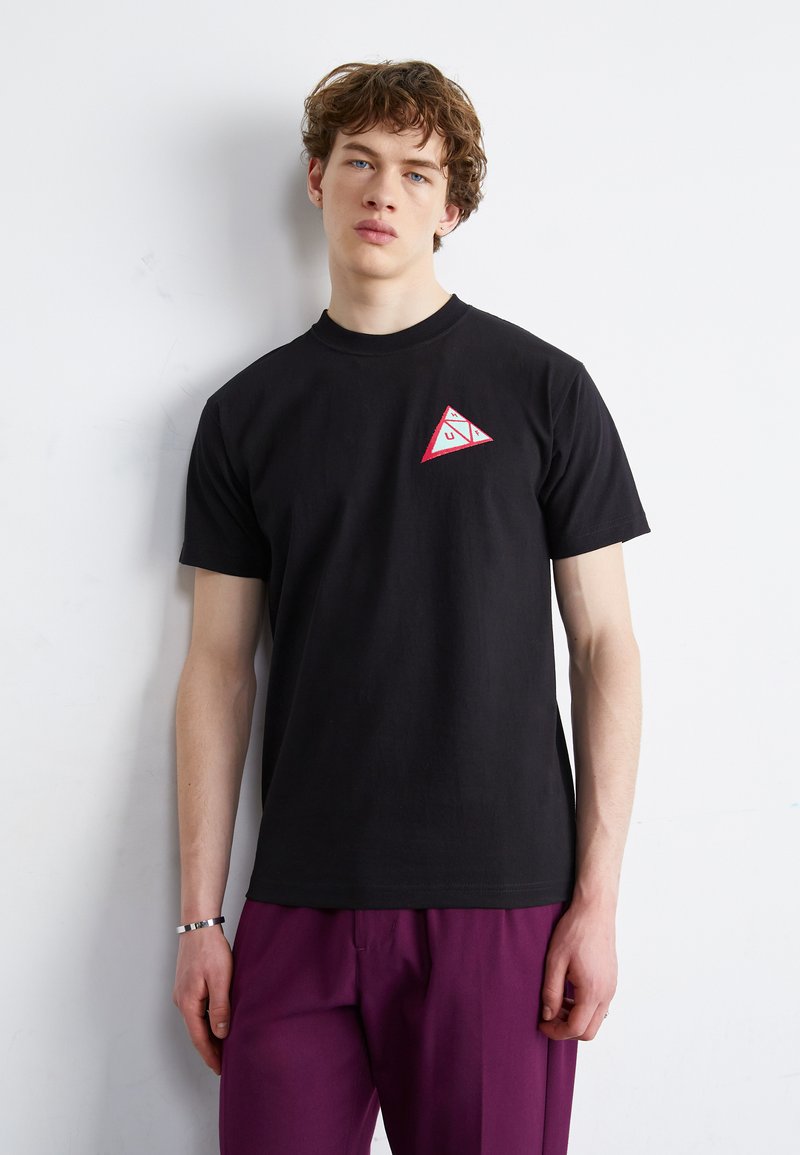 Camiseta de algodón negra que presenta un pequeño logo triangular rojo y blanco en el lado izquierdo del pecho. Tiene mangas cortas y un diseño de cuello redondo.