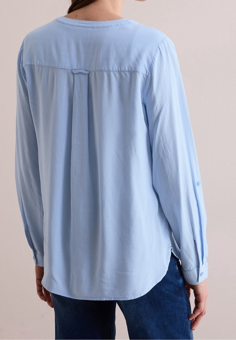 Femme portant un chemisier bleu clair à rayures à manches longues, vue de dos, associée à un jean bleu foncé sur un fond uni.