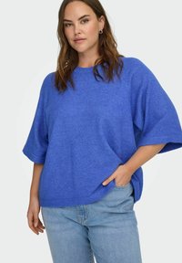 Donna con capelli castani che indossa orecchini a cerchio d'argento, un maglione blu a maniche corte e largo, e jeans azzurri, posando con una mano in tasca.