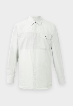 Camicia bianca a maniche lunghe con bottoni, caratterizzata da sottili righe verticali e taschino sul petto a sinistra con un solo bottone.