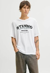 Hvid bomuld t-shirt med sort grafisk tekstdesign, der læser "STUDIO Jack & Jones Siden 1990". Korte ærmer, afslappet pasform, rund hals.