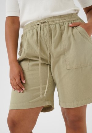 Personne portant un short beige à cordon avec de grandes poches à l'avant et un top blanc, main reposant dans la poche et sur la cuisse.