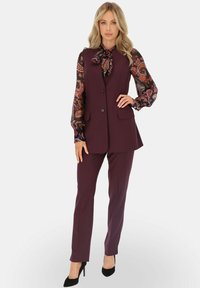 Wino-kolorowy kamizelka o dopasowanym kroju nałożona na wzorzystą bluzkę z długim rękawem z krawatem w kokardę i pasujące dopasowane spodnie, w zestawieniu z czarnymi szpilkami.