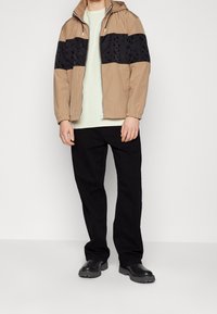 Beżowa kurtka z kapturem, z czarnym wzorzystym panelem na klatce piersiowej, w połączeniu z jasnoniebieską koszulą i czarnymi spodniami. Gładki materiał, luźny krój i zapięcie na zamek.