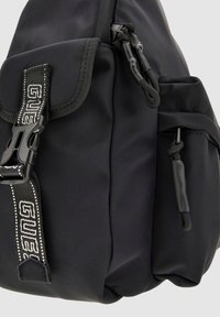 Zaino in nylon nero con chiusura a patta, spallacci regolabili, una tasca laterale e dettagli con logo. Presenta una texture liscia e un design pratico.