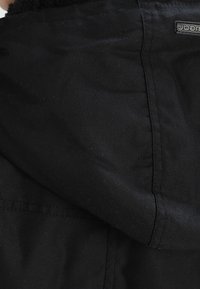 Svart huva jacka med mjukt, texturerat foder. Har en logotagg, slätt tyg och detaljerad sömnad längs sömmarna.