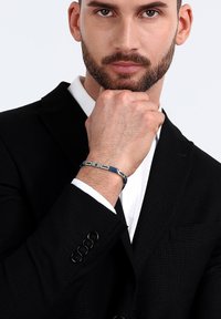 Bracciale in tonalità argento con accenti blu e maglie rettangolari. Indossato a un polso, con giacca nera strutturata e camicia bianca visibili.
