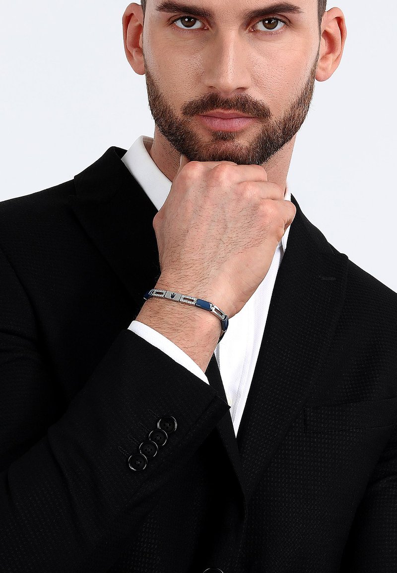 Bracciale in tonalità argento con accenti blu e maglie rettangolari. Indossato a un polso, con giacca nera strutturata e camicia bianca visibili.