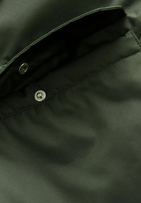 Dunkelgrüner Stoff mit einer glatten Textur verfügt über eine Klappenstasche und einen metallischen Druckknopfverschluss, der die praktischen Designdetails hervorhebt.